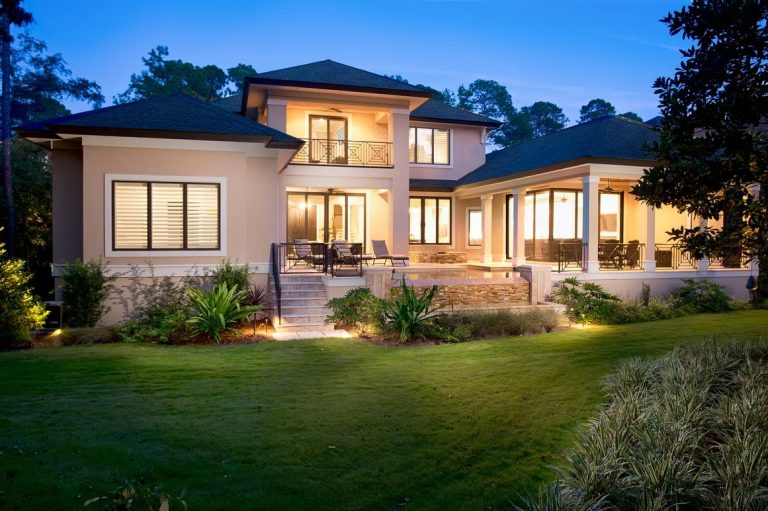 Expansive rear view of a modern luxury estate featuring massive glass windows, a white and charcoal exterior, and a large manicured lawn.